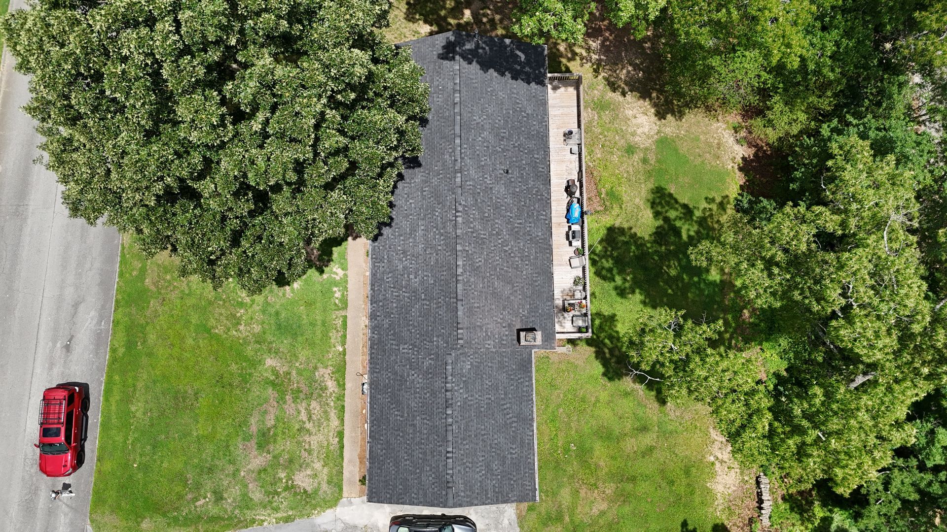 Aerial view of a dark-roofed house beside trees and a grassy yard, with a red car on the road.
