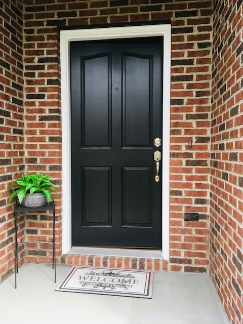 Black front door with brass handle in a brick entryway, with a potted plant and welcome mat.