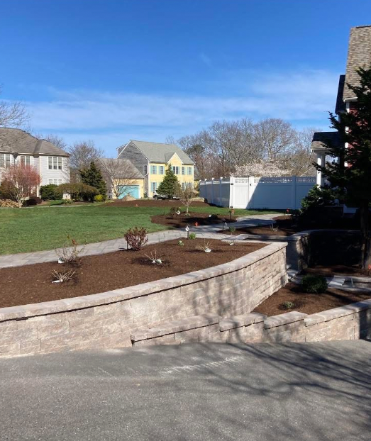 Stone retaining wall with a mulch bed, green lawn, and houses on a sunny day.