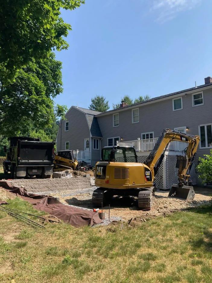 Construction site: Excavator and dump truck clearing dirt near a house with gray siding under a sunny sky.