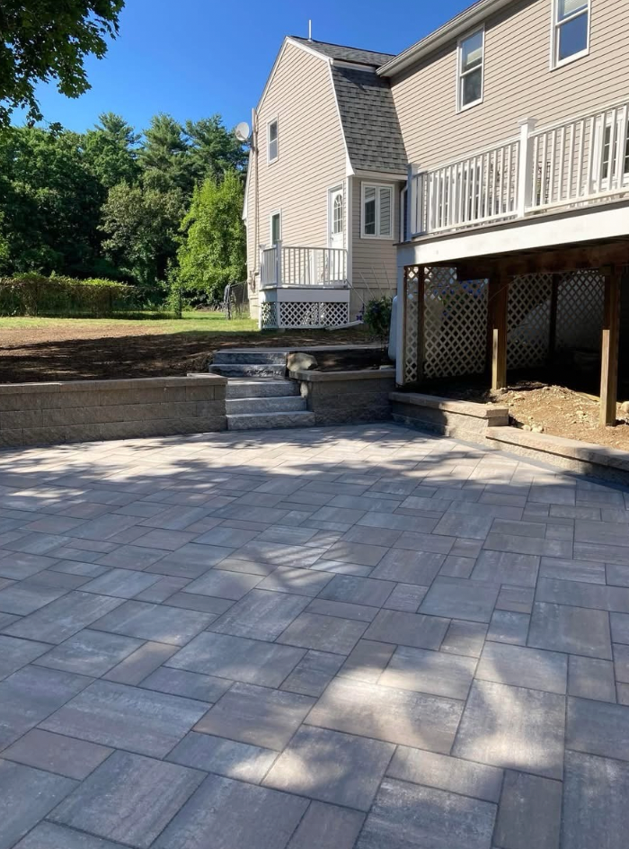 Paver patio with retaining wall, steps, and a two-story beige house with a deck.