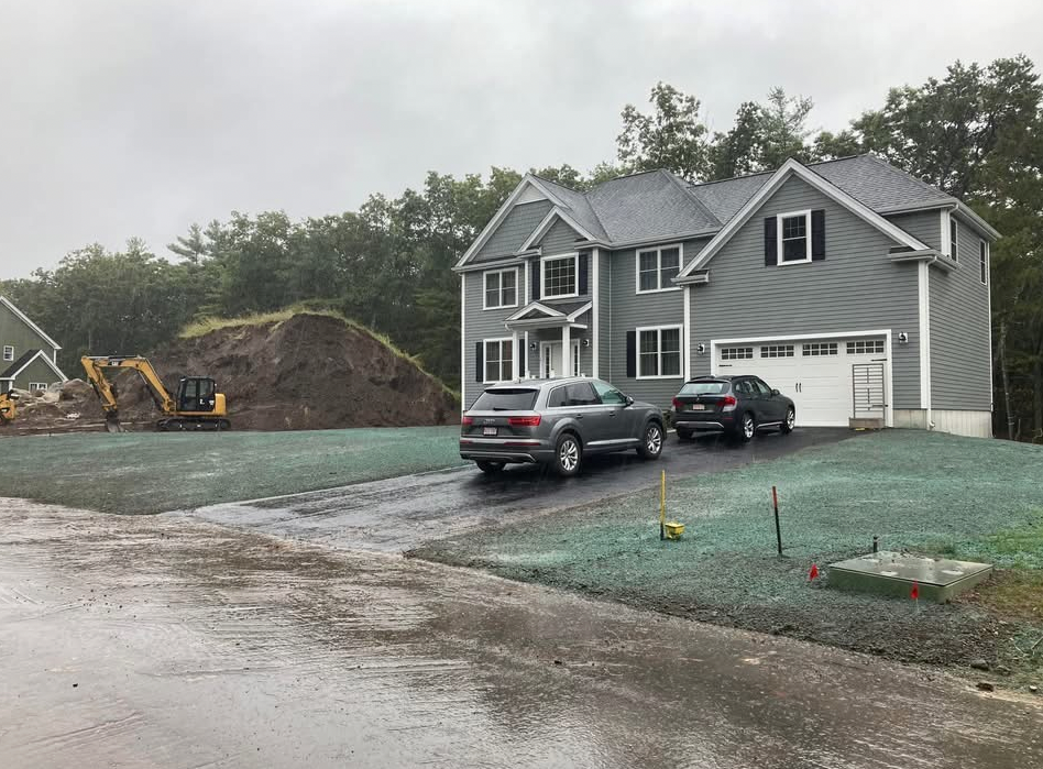 Gray house with cars in driveway; construction equipment and pile of dirt on the left; overcast day.