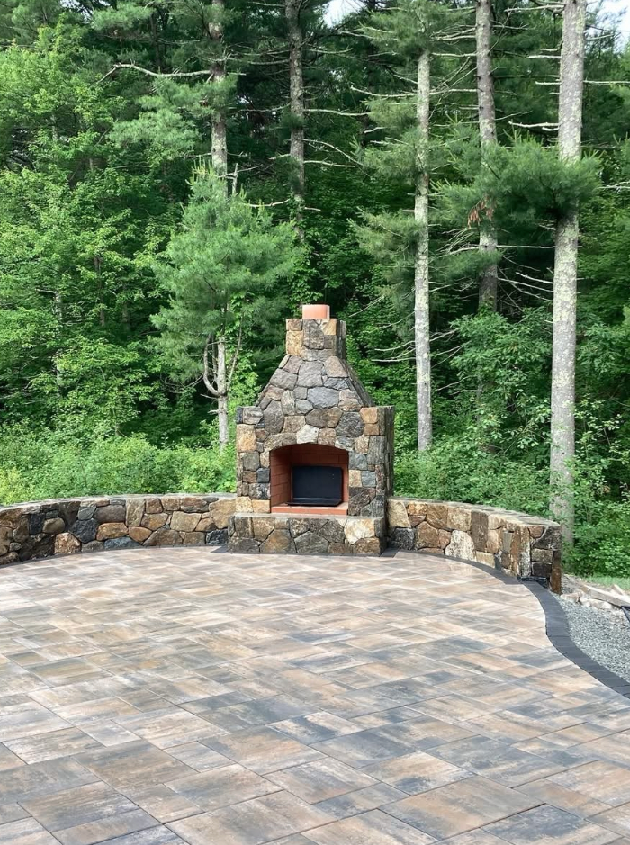 Outdoor stone fireplace on a brick patio with forest backdrop.