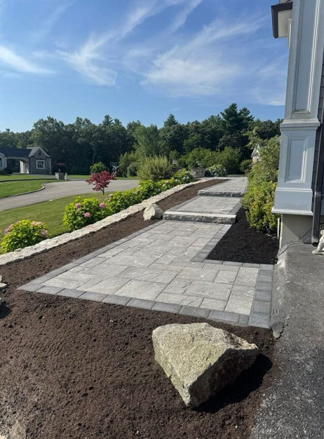 Stone paver walkway with steps leading to a building entrance; landscaping with soil, shrubs, and large rocks.