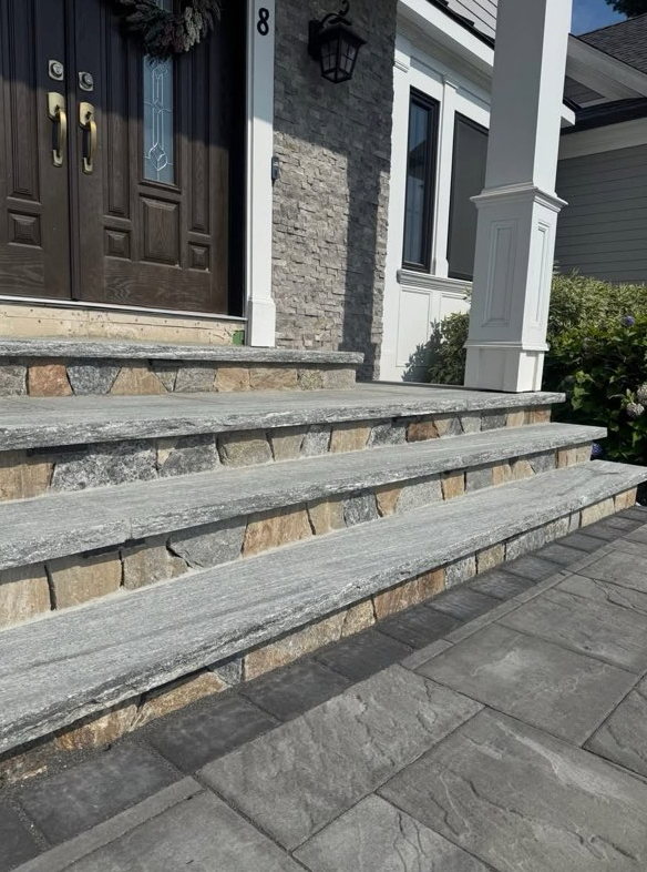 Stone steps leading up to a house with a dark brown door, gray stone facade, and white columns.