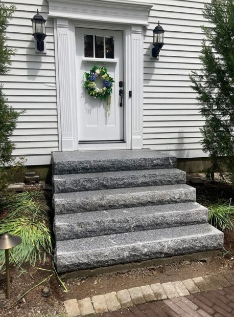 Stone steps leading up to a white front door with a wreath, flanked by two outdoor lights, with white siding.