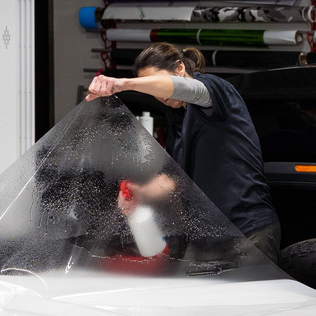 Person spraying water on a large sheet of tint, applying it to a vehicle's surface in a shop setting.