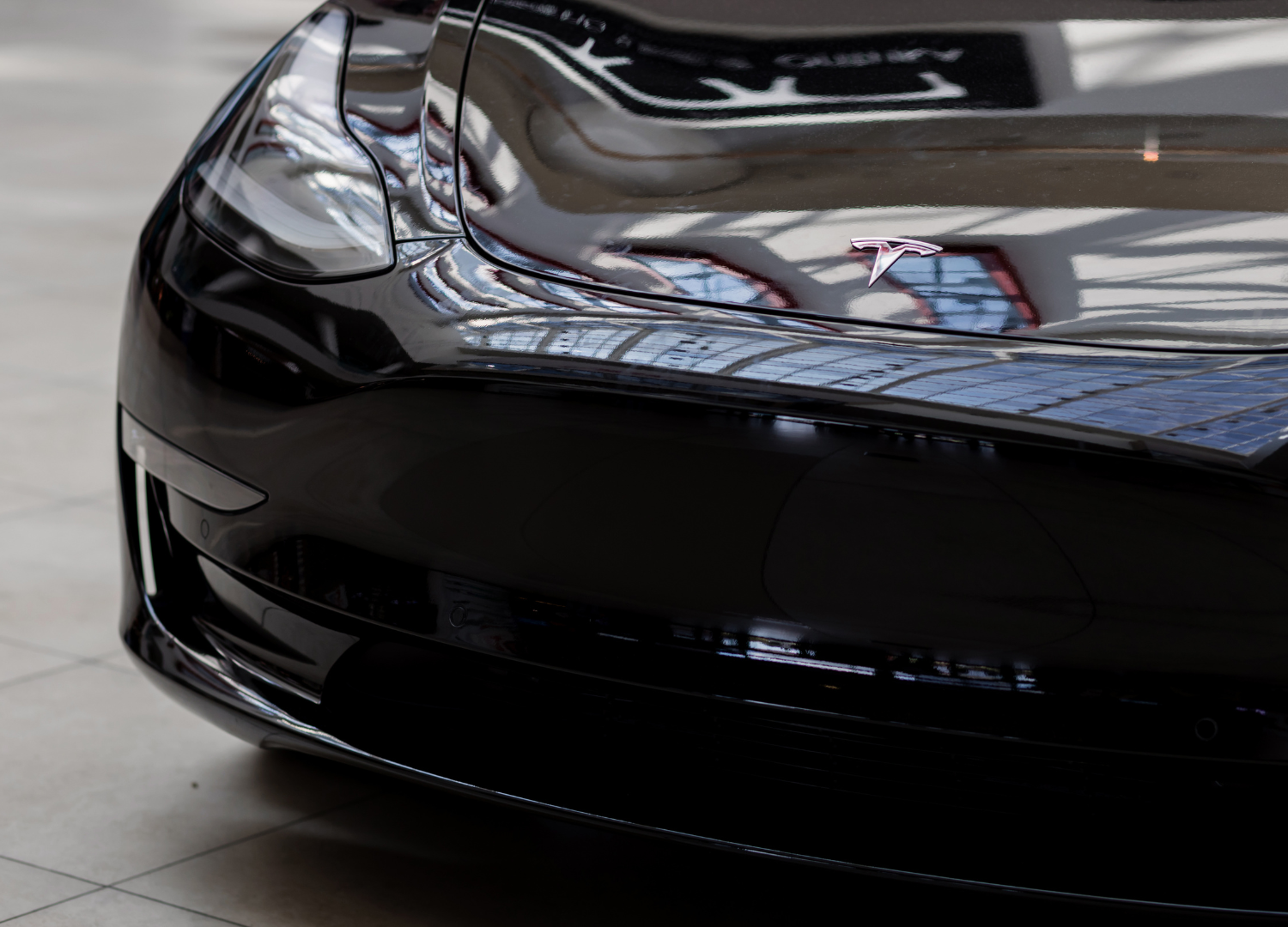 Close-up of a glossy black sports car front bumper and headlight reflecting a showroom ceiling