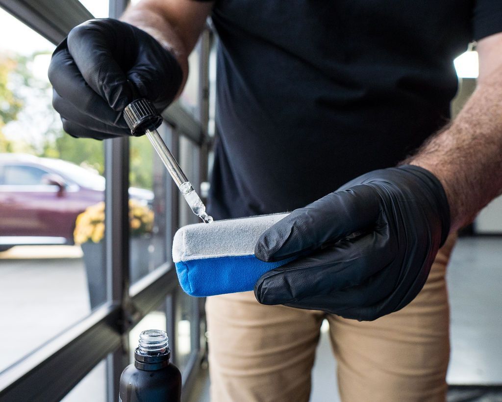 Person in black gloves using a dropper to apply liquid to a blue and gray applicator sponge, near a car.