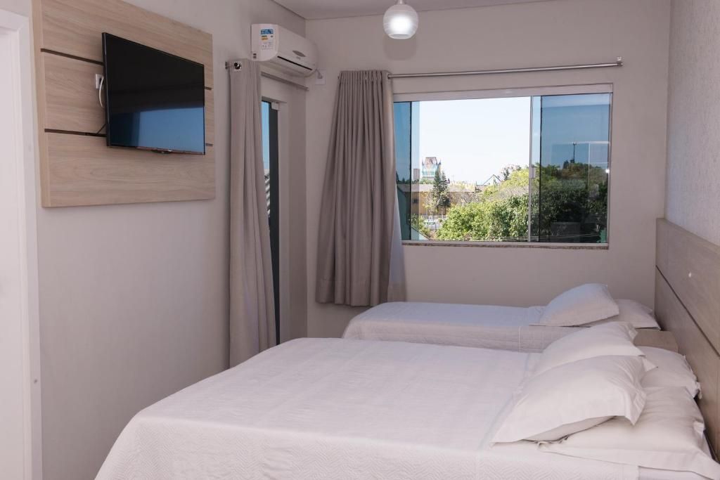 Quarto iluminado com duas camas de solteiro, TV de parede, ar-condicionado e janela com vista para o exterior.