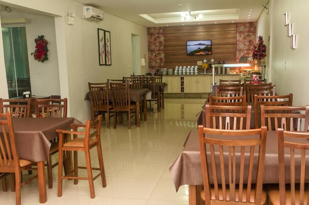 Sala de jantar vazia de um restaurante com mesas e cadeiras de madeira, TV na parede do fundo e piso de azulejo.