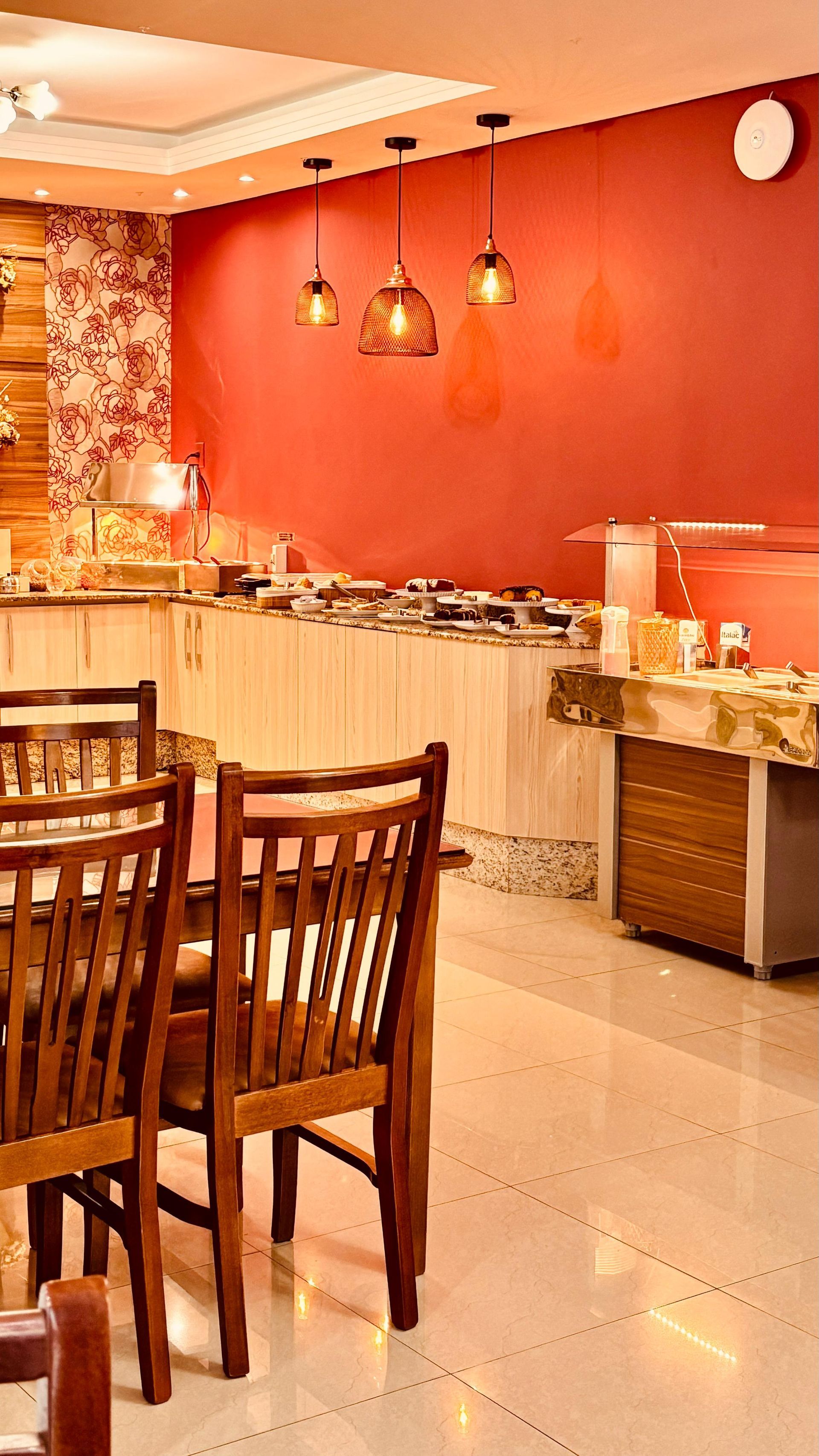 Ambiente acolhedor de restaurante com cadeiras de madeira, paredes vermelhas e luminárias pendentes ao lado de um balcão de buffet.