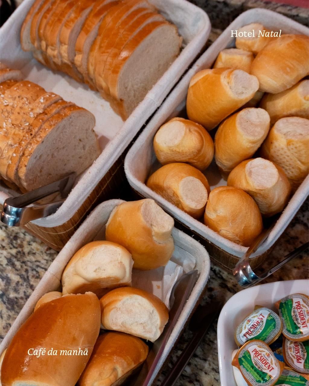 Bandejas com uma variedade de pãezinhos frescos e pães fatiados dispostos sobre um balcão.