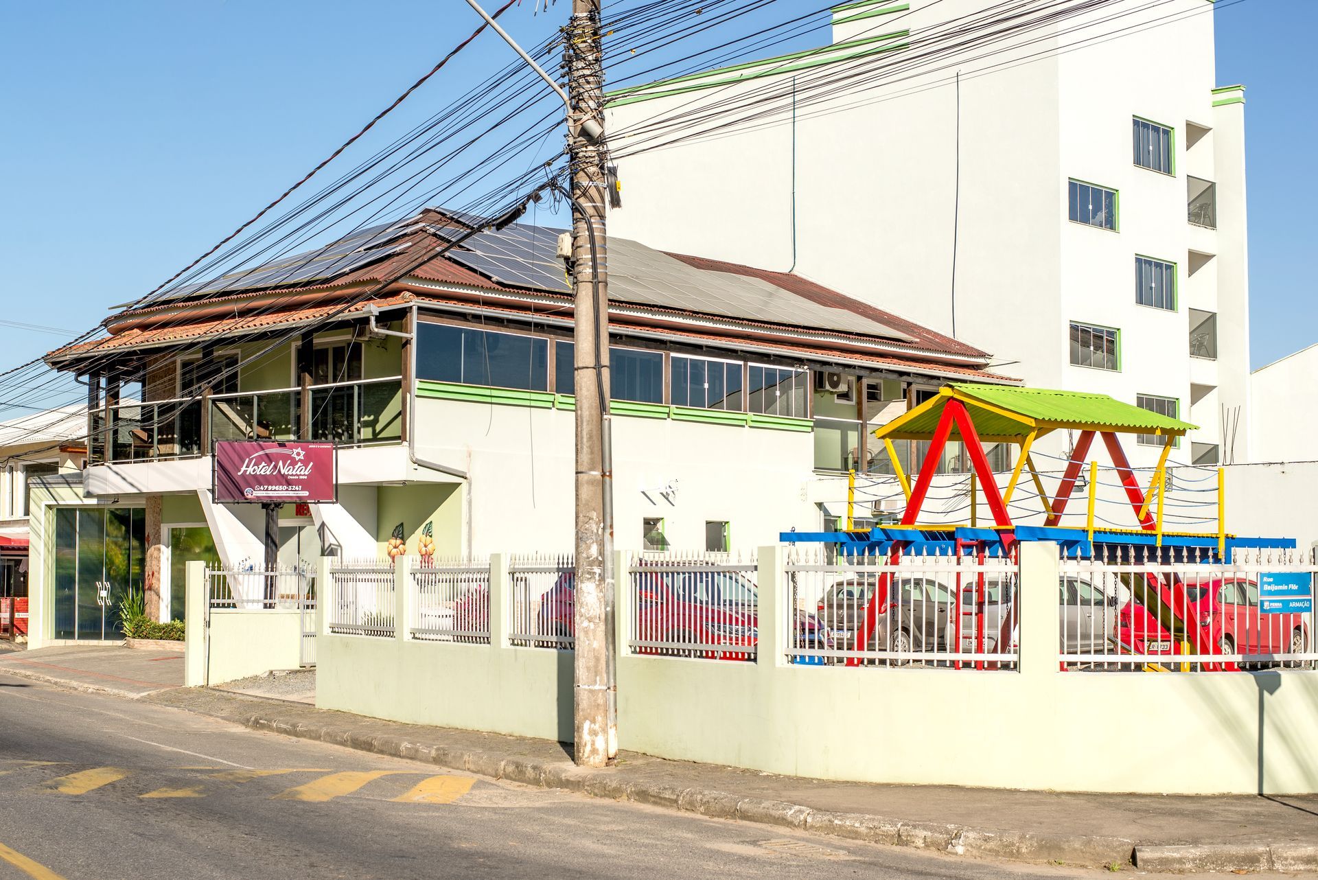 Edifício de rua com um parque infantil cercado e uma estrutura de escalada colorida sob um telhado vermelho.