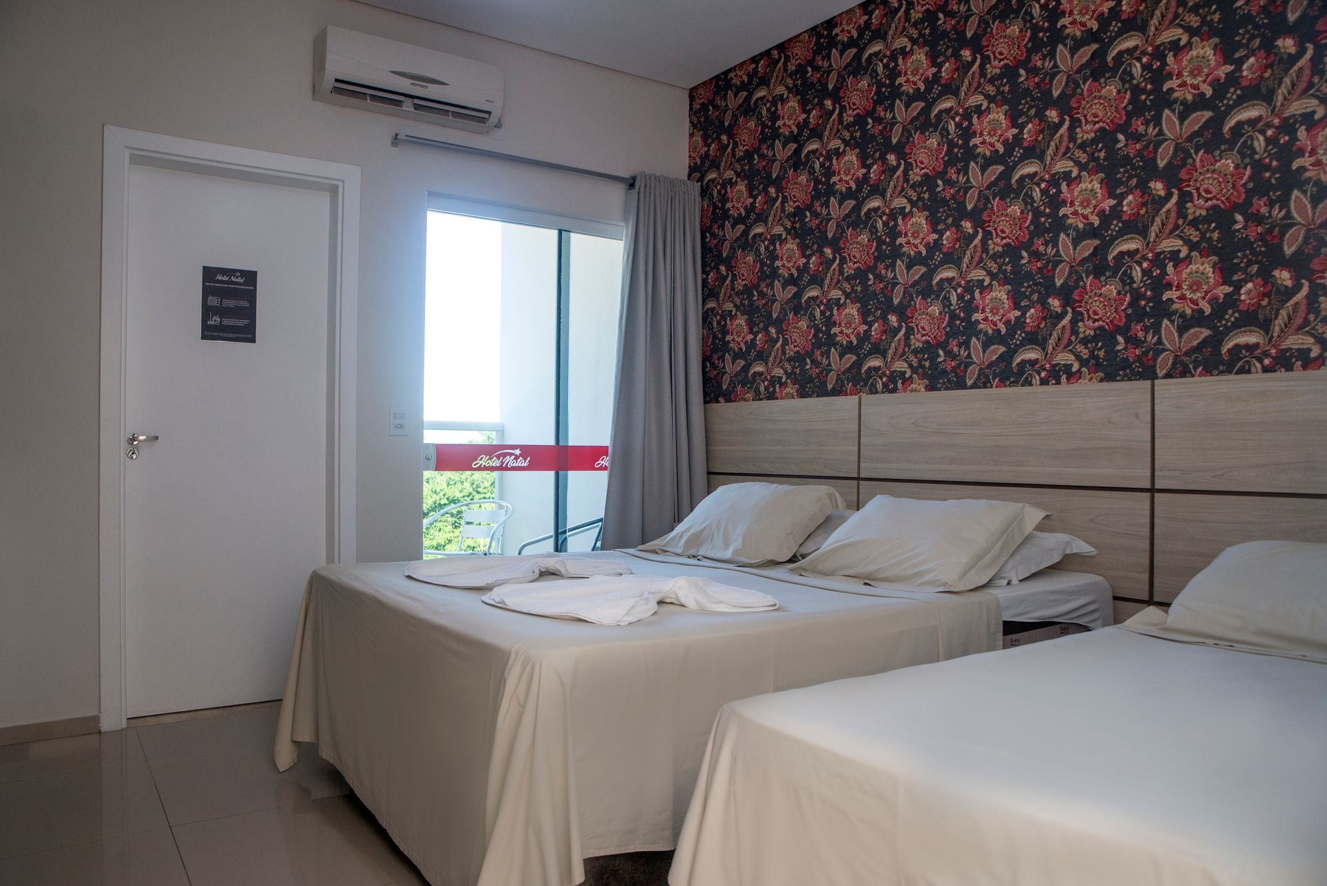 Quarto de hotel pequeno com duas camas, parede com detalhes florais, janela e porta.