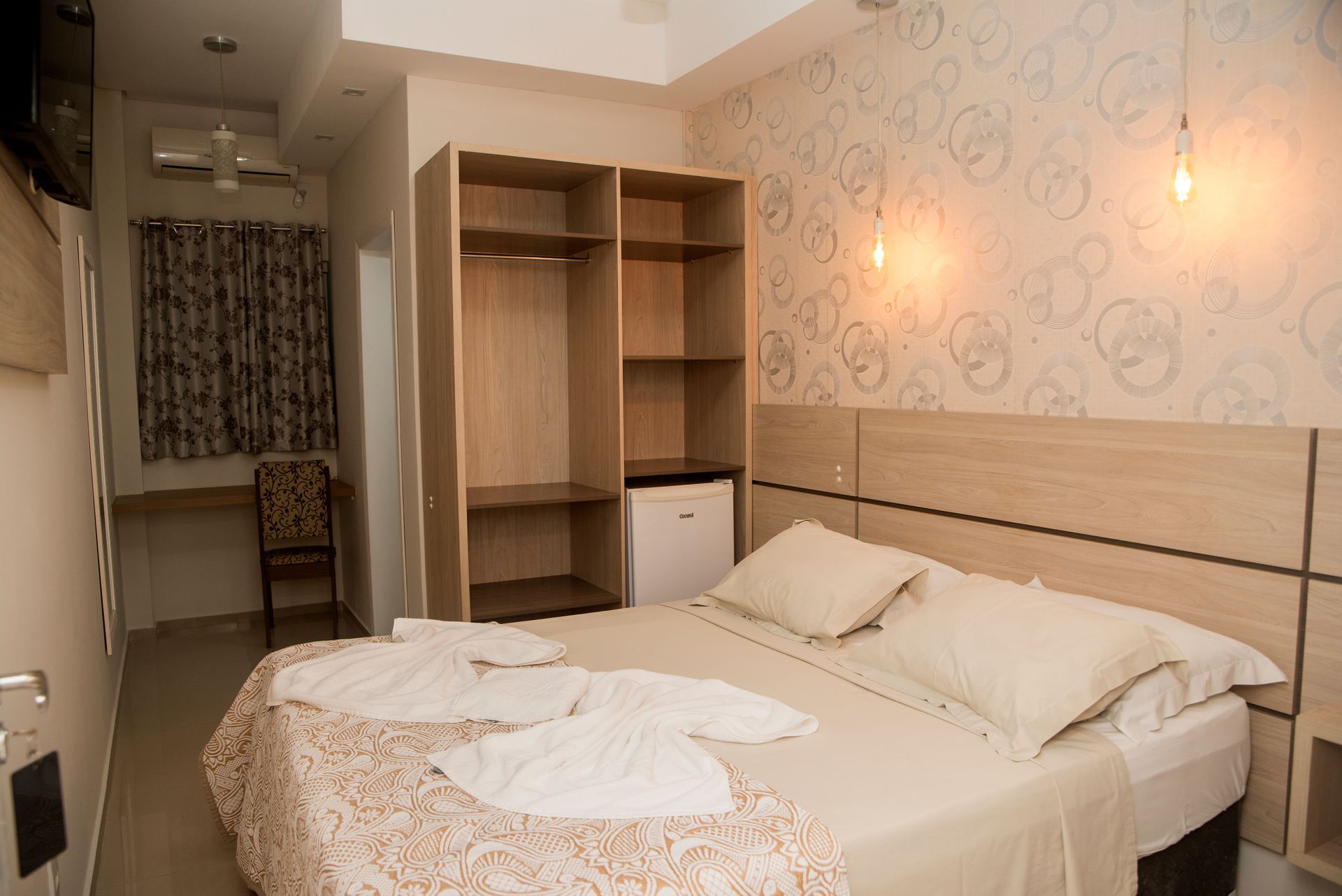 Quarto de hotel pequeno com cama, prateleiras de madeira, papel de parede estampado e porta aberta para um banheiro.