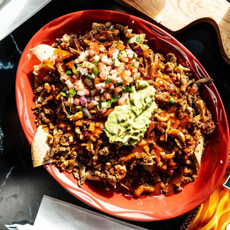 Red bowl of loaded nachos topped with guacamole and diced onions on a dark table