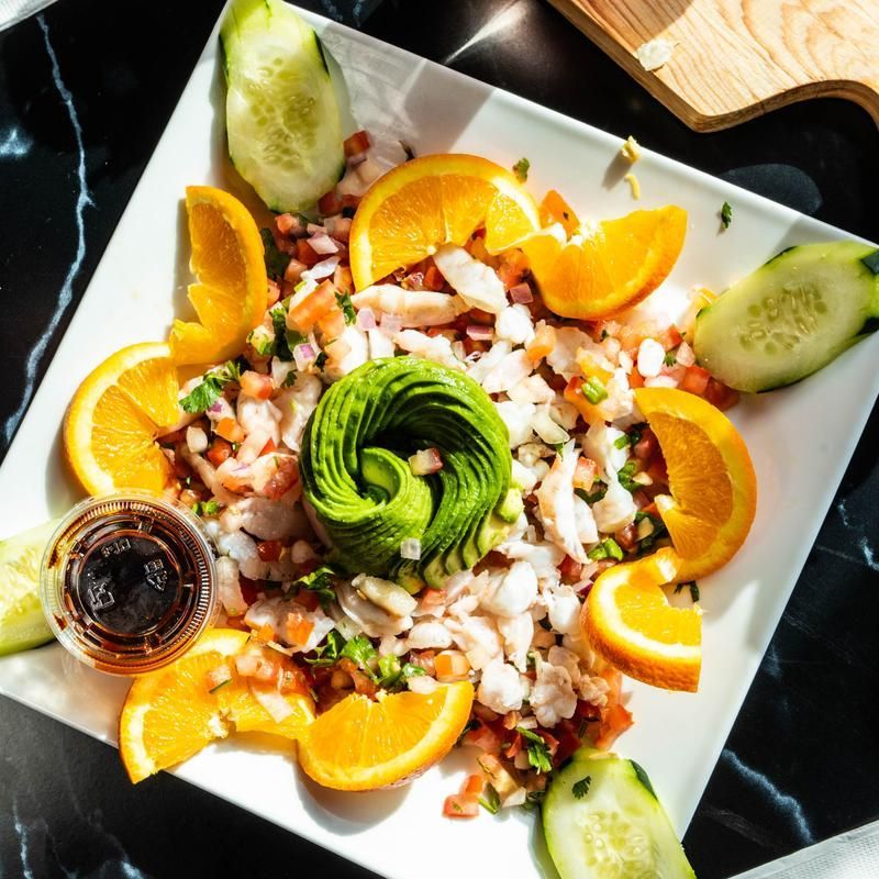 Ceviche on a white plate with avocado garnish, orange slices, cucumber, and a small sauce cup