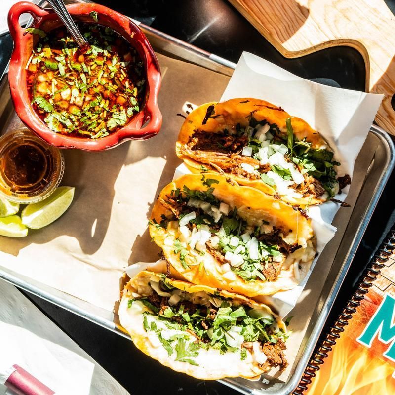 Tray of tacos with salsa, lime wedges, and a drink on a table.