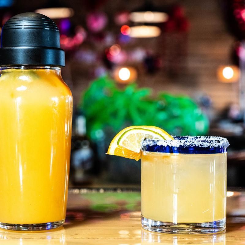 Two yellow cocktails in glassware, one with a lemon slice and blue salted rim, on a bar counter
