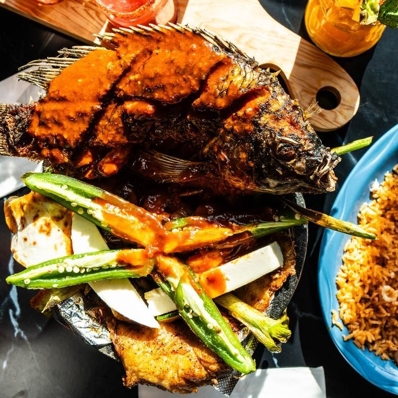 Grilled whole fish with green beans and rice on a black plate, with sauce and drinks nearby