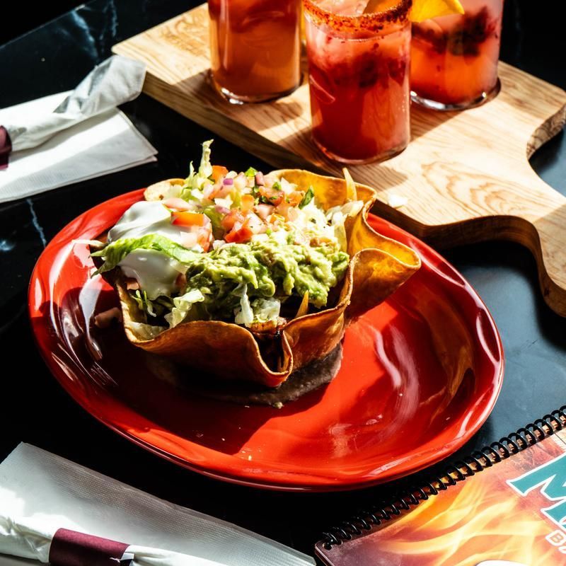Fresh salad in a crisp tortilla bowl on a red plate, with drinks on a wooden tray nearby.