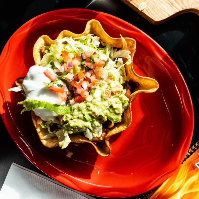 Waffle bowl taco on a red plate, topped with guacamole, lettuce, sour cream, and diced tomatoes.