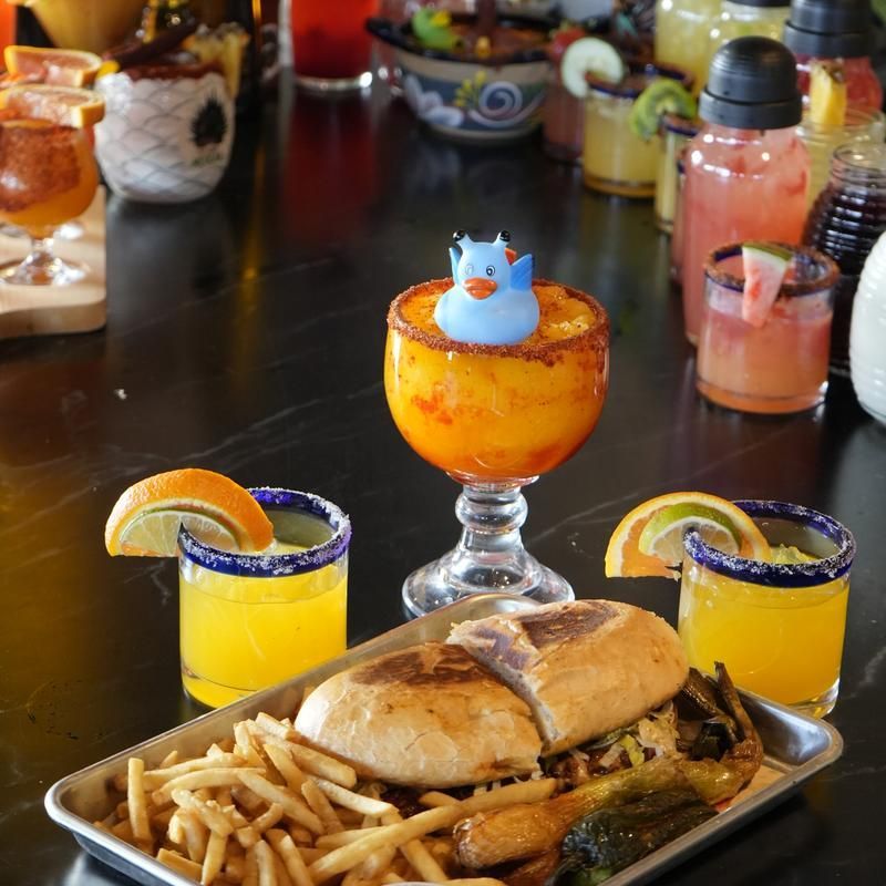 Colorful restaurant meal with fries, sandwiches, cocktails, and a small blue toy bird centerpiece