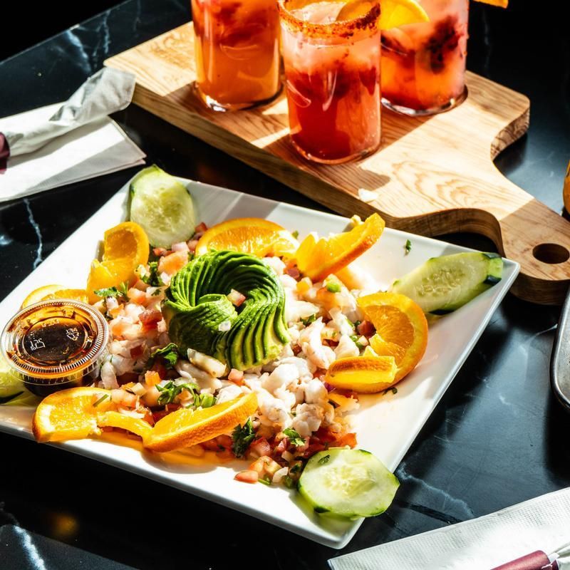Colorful ceviche plated with avocado and citrus wedges beside three iced drinks on a wooden tray