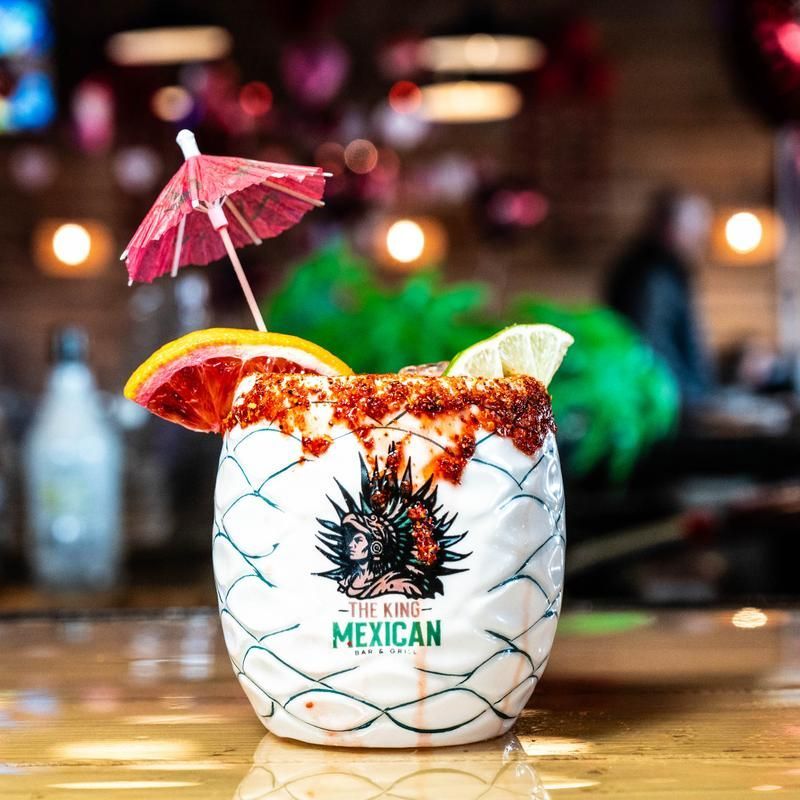 Colorful cocktail with lime, orange, and paper umbrella in a branded Mexican glass on a bar