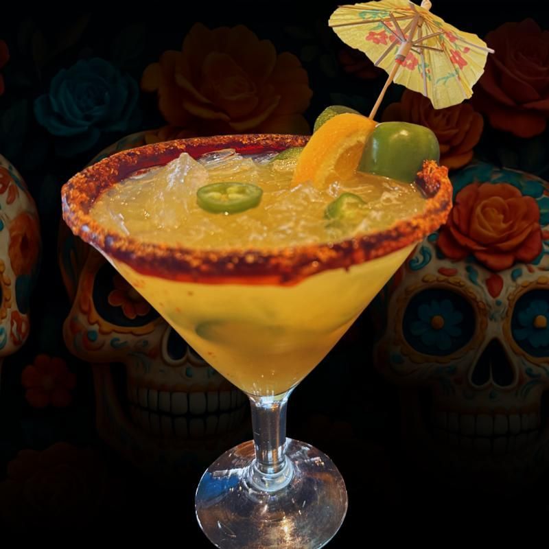 Yellow cocktail in a martini glass with lime garnish and paper umbrella, against a Day of the Dead skull backdrop