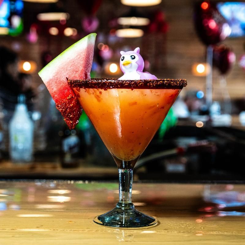 Orange cocktail in a martini glass with a watermelon wedge and small owl garnish on a bar counter