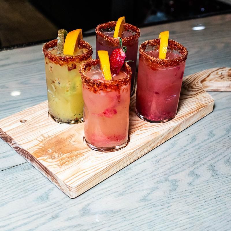 Four colorful salted cocktails with fruit garnish on a wooden serving board