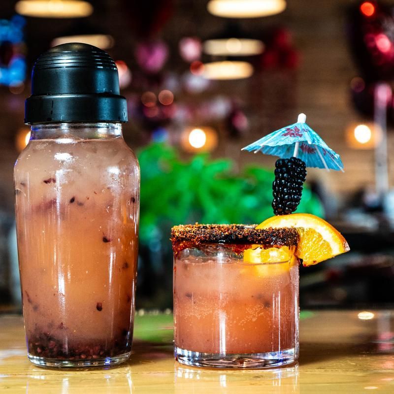 Two tropical cocktails on a bar, one in a bottle and one garnished with an orange slice and tiny umbrella