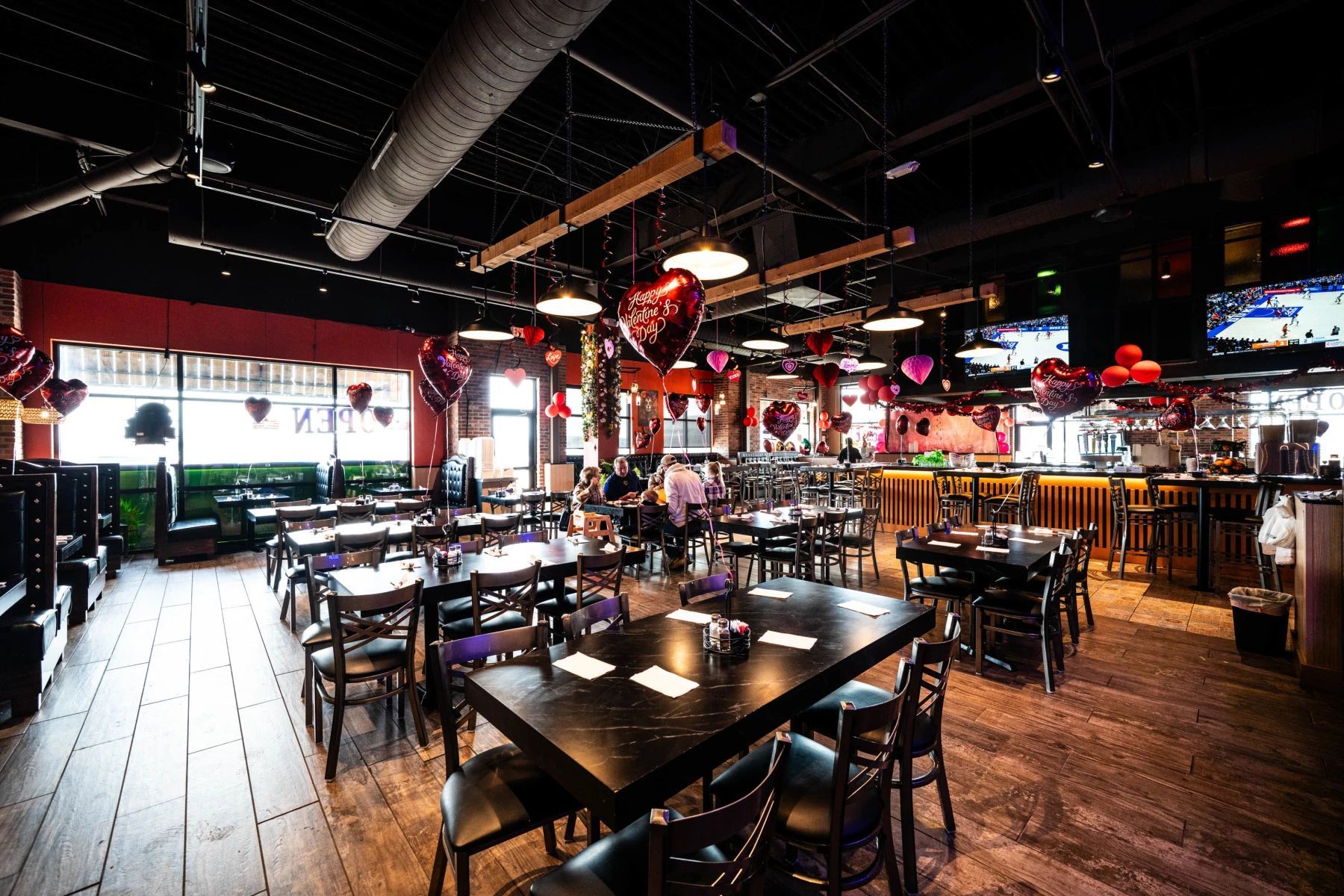 Empty restaurant dining area with black tables and chairs under red hanging lights and a wood bar.