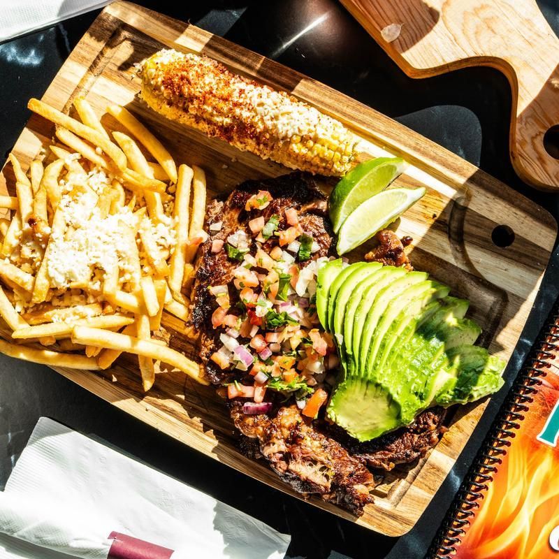 Grilled steak with avocado, corn, fries, and salsa on a wooden tray under bright sunlight