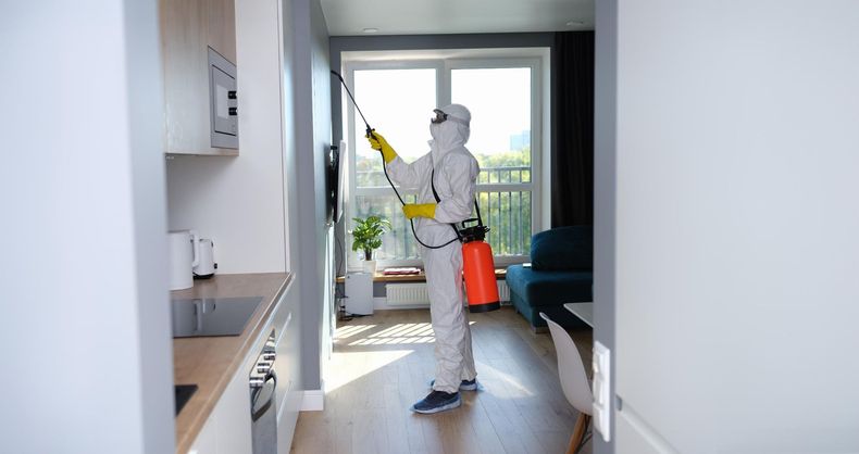 Person in white coveralls cleaning a window with a yellow spray bottle in a bright apartment.