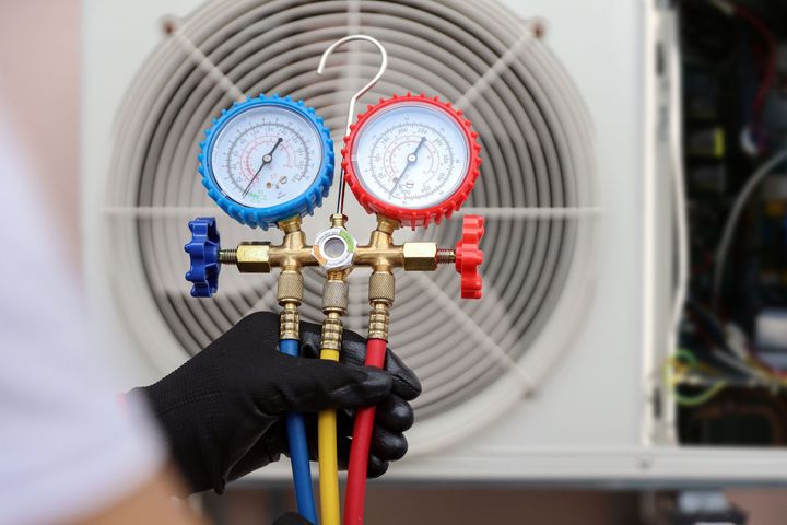 Gloved hand holding manifold gauge set connected to AC unit, checking pressure levels.