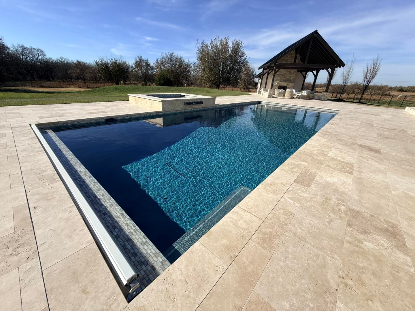 Empty outdoor swimming pool with blue tile, concrete deck, and a small pavilion nearby