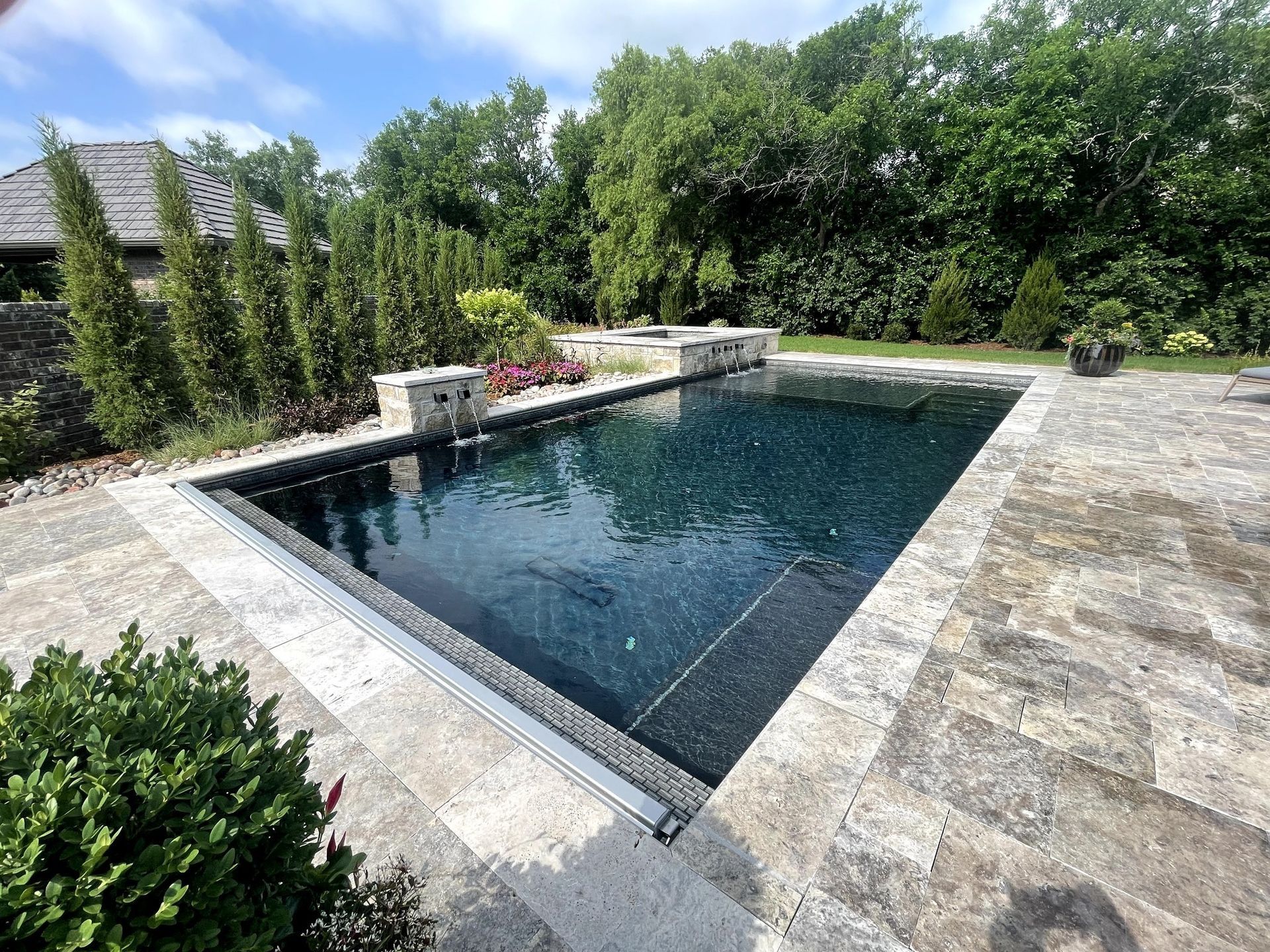 Backyard stone patio with a rectangular swimming pool surrounded by trees and shrubs