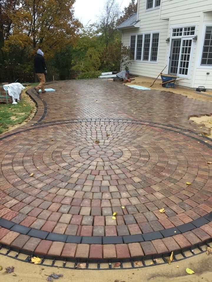 Brick-and-stone patio under construction beside a house, with a circular pattern and workers nearby