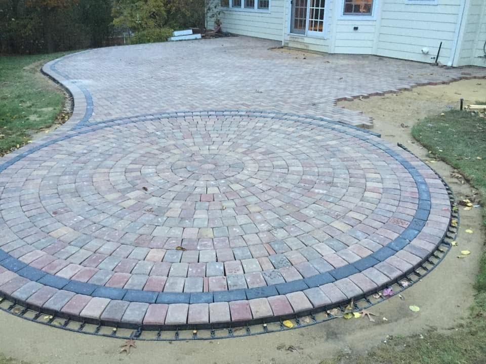 Circular brick paver patio beside a house, with a gray border and curved walkway extension.