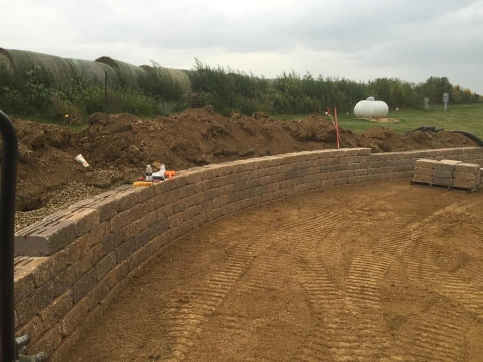 Curved brick retaining wall under construction beside a dirt hillside and grassy field