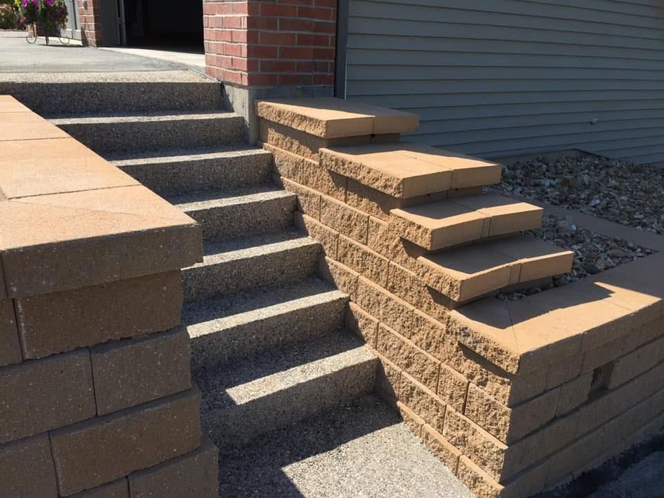 Stone steps beside a brick house and retaining wall, with sunlit concrete and gravel landscaping