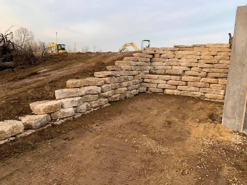 Stone retaining wall under construction on a dirt hillside, with excavators in the background.