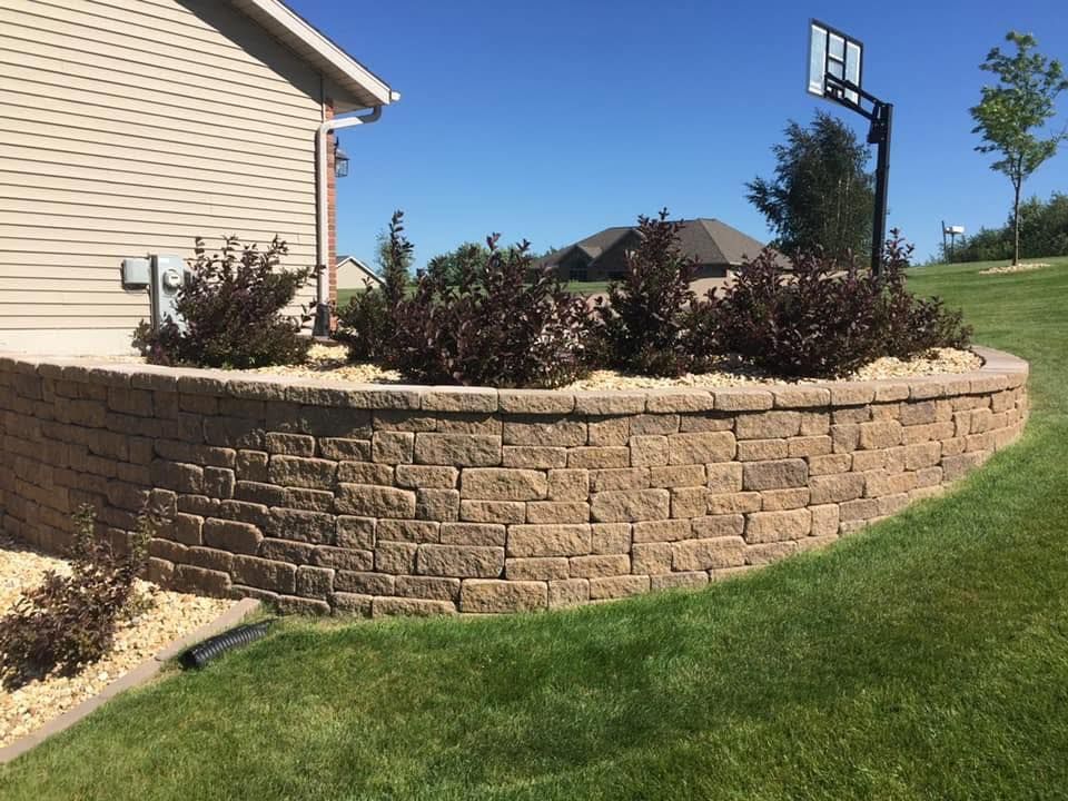 Curved stone retaining wall beside a house, with shrubs, green lawn, and a basketball hoop nearby.
