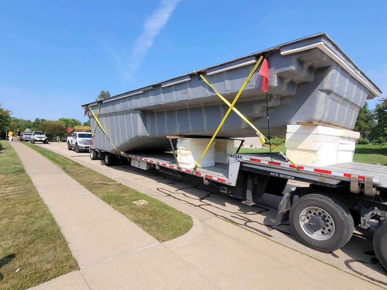 Large metal container secured on a flatbed trailer with yellow straps, parked beside a sidewalk.