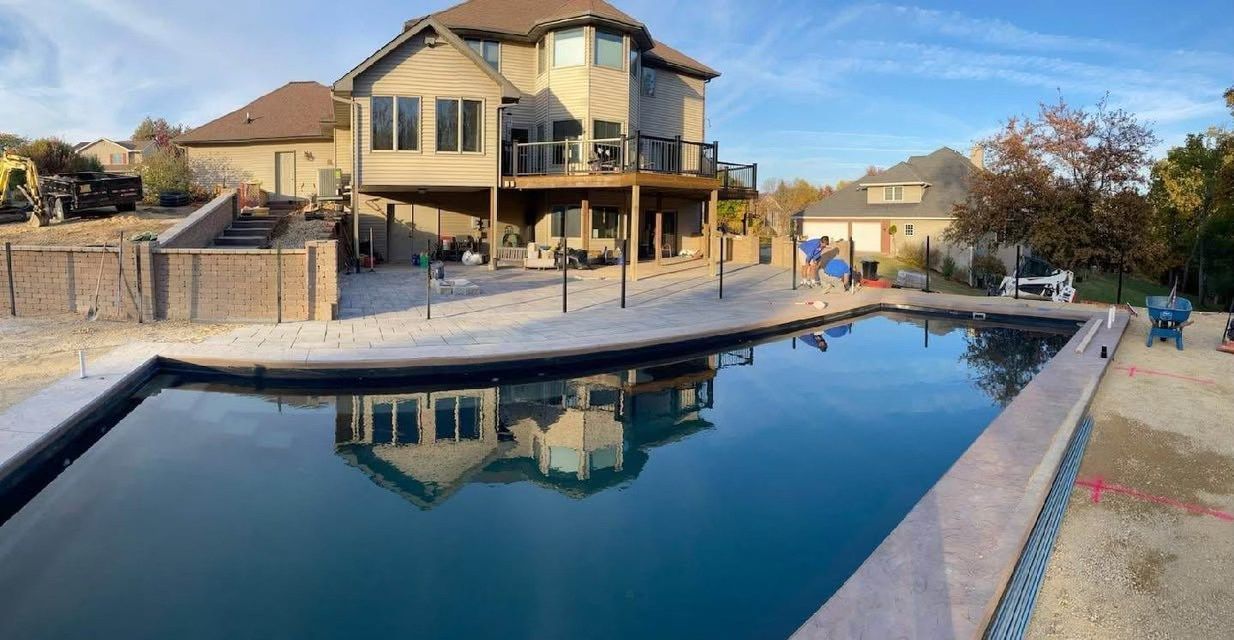 Backyard pool reflecting a tan two-story house under a blue sky, with stone deck and fence