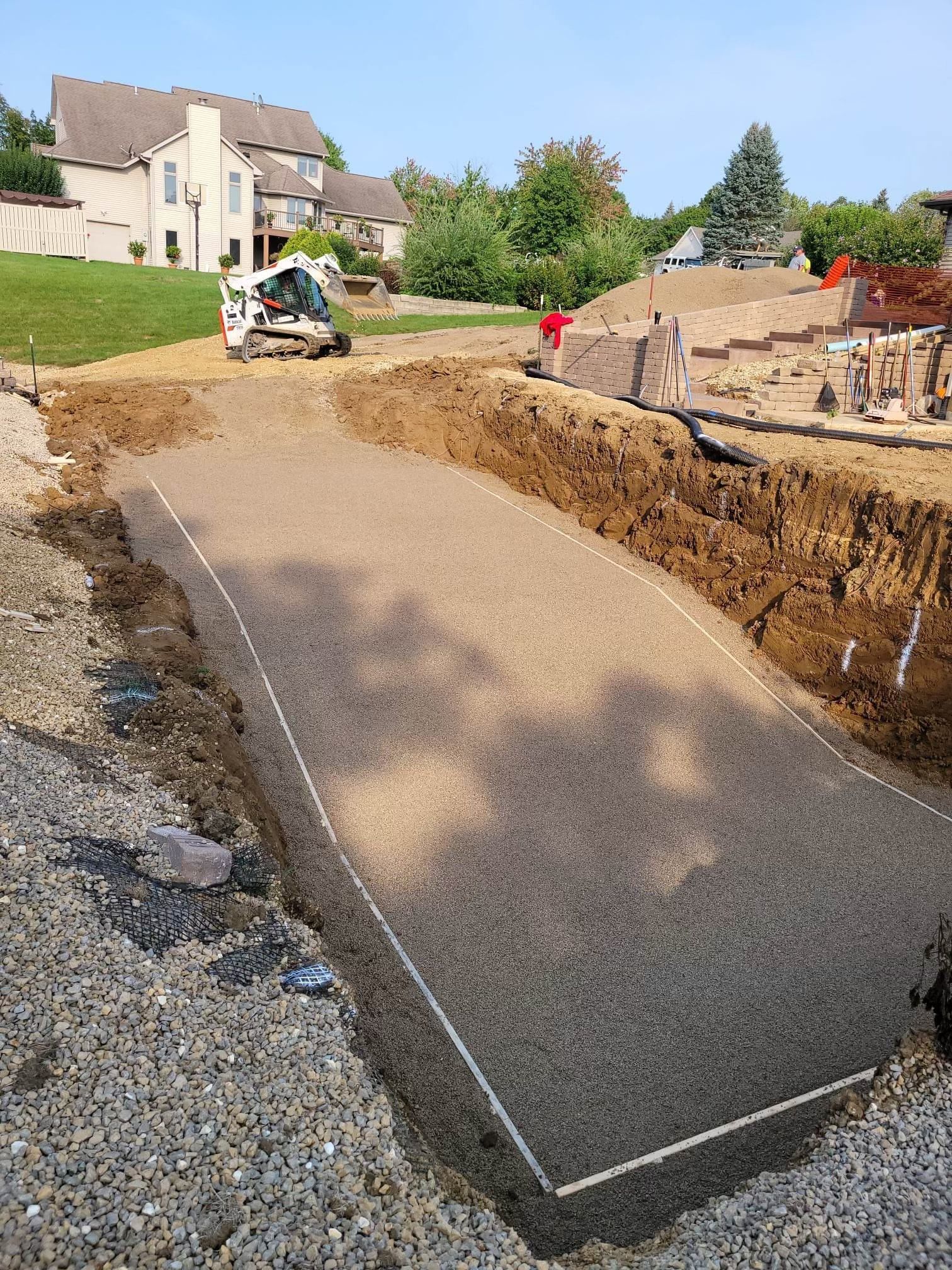 Construction site with excavator shaping a gravel path beside a trench and houses