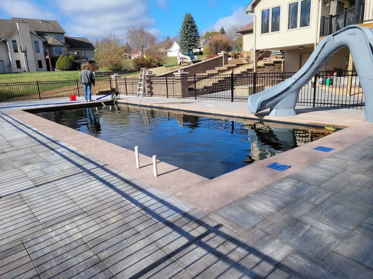 Outdoor pool with a slide, surrounded by stone deck and houses under a clear sky